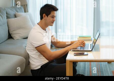An Asian man working at home, chatting online with a colleague on his laptop while on the couch in living room at home. Stock Photo