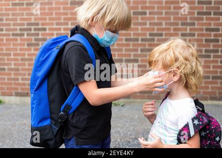 Kids in masks. Back to school concept after reopening. Schoolboy putting mask on his little sister during corona virus outbreak. Small kids wear backp Stock Photo
