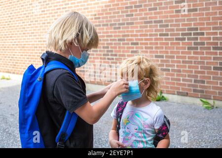 Kids in masks. Back to school concept after reopening. Schoolboy putting mask on his little sister during corona virus outbreak. Small kids wear backp Stock Photo