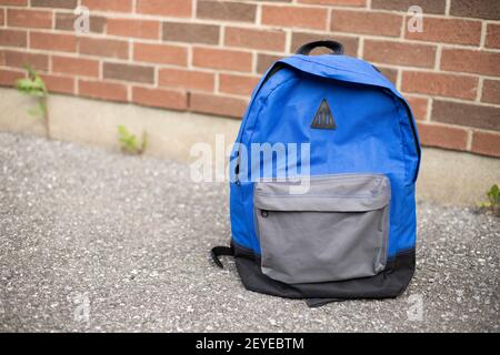School backpack on the road. Blue boy's backpack on the pavement near red brick wall of school in the background. Back to school educational concept. Stock Photo
