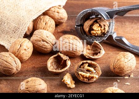 Angled selective focus image of walnuts in shell scattered out of a vintage burlap sack onto wooden table. Some are cracked using metal walnut pliers Stock Photo