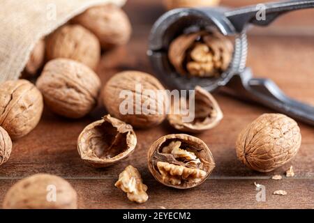 Angled selective focus image of walnuts in shell scattered out of a vintage burlap sack onto wooden table. Some are cracked using metal walnut pliers Stock Photo