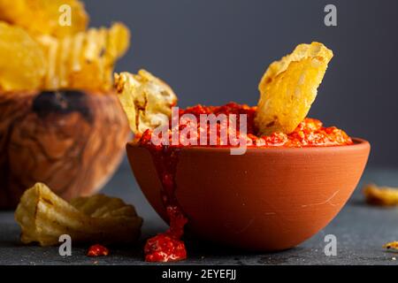 A wooden bowl of potato chips with a terra cotta bowl of salsa or hot pepper dipping sauce. A chips has been dipped in. Some mess and spilling on dark Stock Photo