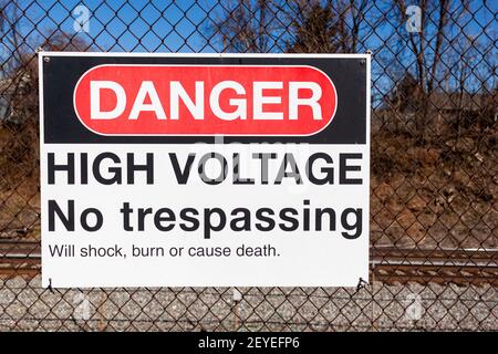 Danger, high voltage, no trespassing sign on a fence in front of a rail road. It lists possible health hazards including shock, trauma and death under Stock Photo