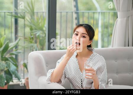 Smiling healthy young woman holding pill glass of water sit on sofa at home Stock Photo