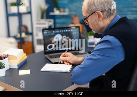 Smart thoughtful technician writing on notebook. Experienced engineer working from home office on mechanical project using laptop computer technology and wife is reading a book on sofa. Stock Photo