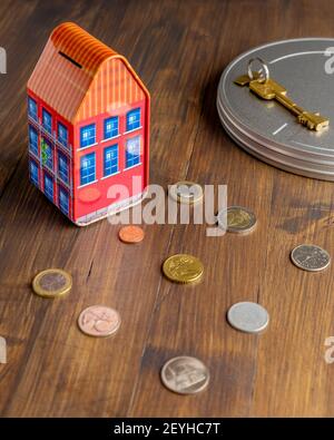 A colorful little house shaped piggy bank on a wooden table with coins and a safe key Stock Photo