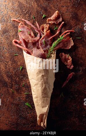 Slices of spicy dry-cured meat with rosemary in a paper bag on an old brown background. Top view. Stock Photo