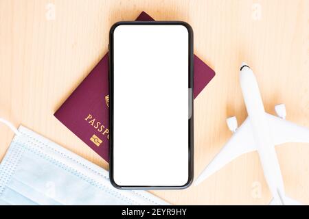 Flat lay with copy space, a model aircraft, passport, face mask and a smartphone with white screen are arranged on a wooden background. Stock Photo