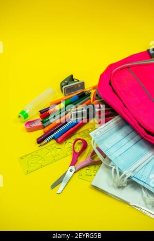 School supplies like pencil scissor eraser ruler watercolor mask alcohol sanitizer inside a pink bag backpack on yellow background. Back to school con Stock Photo