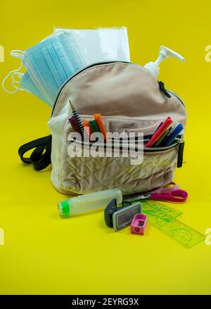 School supplies like pencil scissor eraser ruler watercolor mask alcohol sanitizer inside a brown bag backpack on yellow background. Back to school co Stock Photo