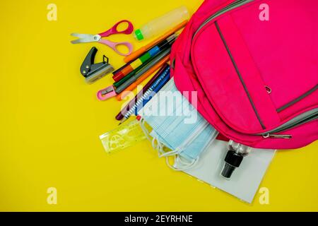 School supplies like pencil scissor eraser ruler watercolor mask alcohol sanitizer inside a pink bag backpack on yellow background. Back to school con Stock Photo