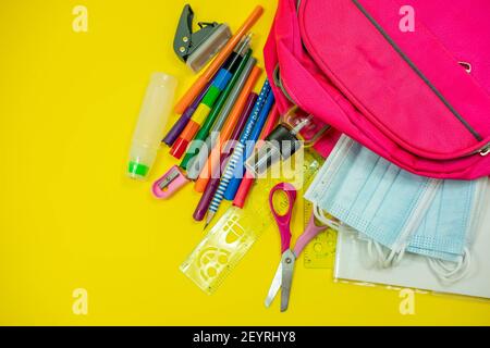 School supplies like pencil scissor eraser ruler watercolor mask alcohol sanitizer inside a pink bag backpack on yellow background. Back to school con Stock Photo
