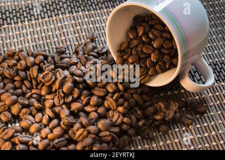 A rose coffee cup full of coffee beans spilling out on a bamboo mat background Stock Photo