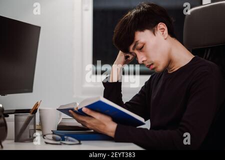 Young handsome asian man sleeping from heavy reading book at work desk late at night, Knowledge and learning concept. Stock Photo