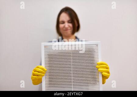 Woman holds ventilation grill with dust filter to clean it. Dirty and dusty white plastic, harmful for health. Cleaning lady with disguised expression Stock Photo