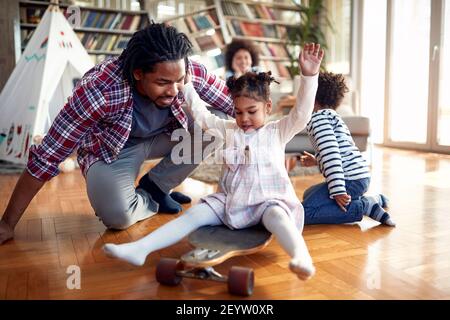 A happy father riding her daughter on the skateboard in a cheerful atmosphere at home. Family, together, love, playtime Stock Photo