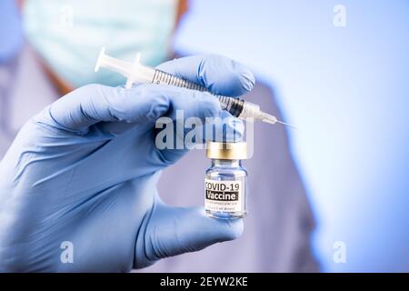 Close-up of hand with surgical glove holding covid-19 vaccine vial and syringe by Asian medical doctor in background Stock Photo
