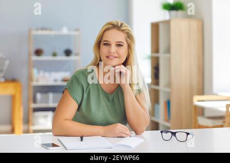 Young smiling blonde woman office worker sitting in office with notebook and pen Stock Photo