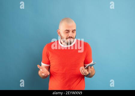 A bald middle-aged man with a beard and a red T-shirt on a blue background, fearfully, covers his face with his hands Stock Photo