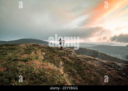Photographer on top summit of mountain hill at dawn as sunrise illuminates clouds in sky beautiful. Using a tripod in rural landscape Peak District Stock Photo