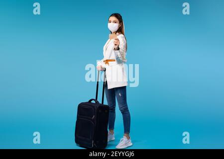 Young caucasian woman in medical mask with suitcase looking to the camera while standing near modern gates on railroad station, showing bank card Stock Photo