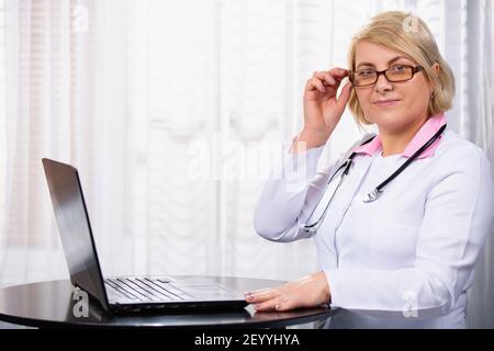 Serious female doctor using laptop and typing notes in medical journal sitting at desk. Senior woman,professional medic physician wearing white coat Stock Photo
