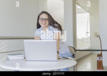 A cheerful Hispanic woman working at home with a laptop and books on a table from home during a quarantine Stock Photo