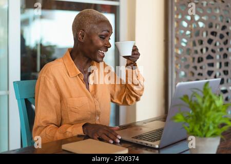 Afro senior woman having fun doing video call using computer while drinking coffee in bar restaurant Stock Photo