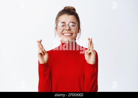 Young female employee in glasses making a wish, cross fingers for good luck and close eyes, pleading god dream come true, standing excited against Stock Photo