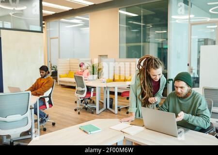 Two young contemporary office managers in casualwear looking at laptop display while discussing some points of business presentation Stock Photo