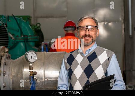 Smiling Manager With Digital Tablet Looking at Camera. Engineer Standing in District Heating Power Plant. Digital Technology Concept. Industry 4.0 Stock Photo