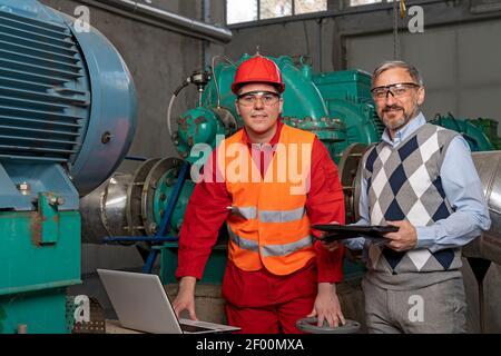 Young Worker Using Laptop Computer for Data Checking. Portrait of Manager and Worker in District Heating Power Plant. Teamwork Concept. Industry 4.0 Stock Photo