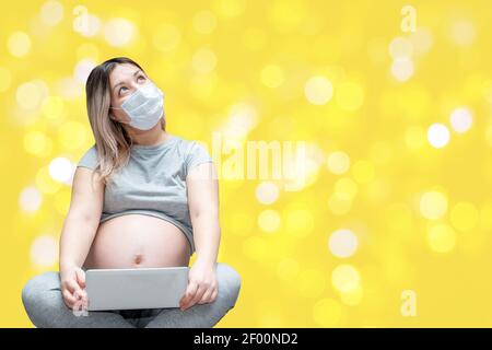 Young female in expectancy wearing protective face mask and looking up in thoughts of childbirth while using tablet on yellow background with bokeh Stock Photo