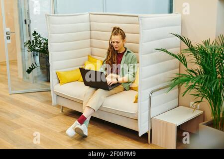 Confident young female office manager in casualwear looking at laptop display while sitting on soft couch and preparing presentation Stock Photo