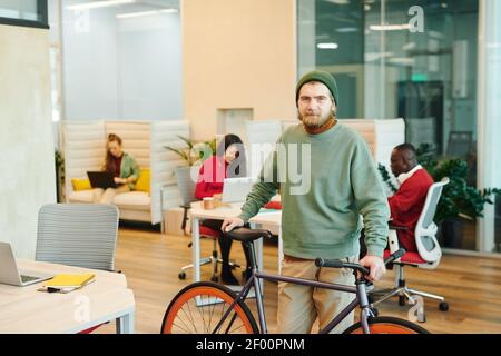 Bearded male office manager in casualwear looking at you while standing by his workplace and holding bicycle against working colleagues Stock Photo