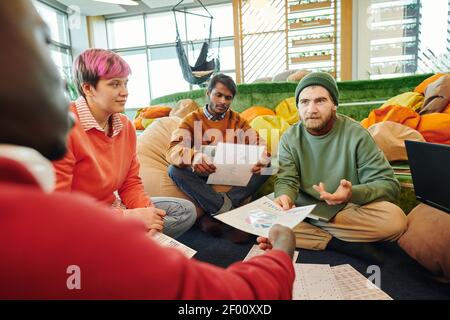 Group of young intercultural financial managers in casualwear discussing papers with diagrams and charts at working meeting on the floor Stock Photo