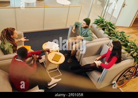 Group of young contemporary intercultural colleagues sitting in armchairs opposite each other and discussing ideas for new business project Stock Photo