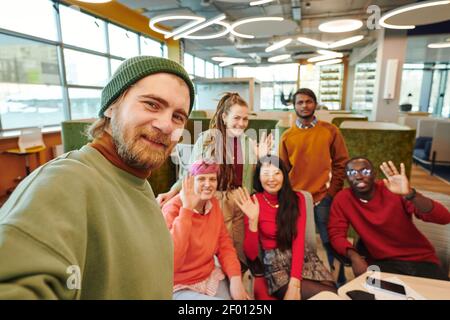 Happy young bearded businessman in casualwear making selfie with his cheerful intercultural colleagues sitting by workplace on background Stock Photo