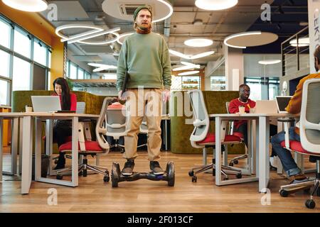 Young contemporary businessman on gyroscope moving along large open space office between desks where his colleagues using computers Stock Photo