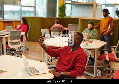 Happy young African businessman in casualwear and eyeglasses making selfie or video call while sitting by workplace against colleagues Stock Photo