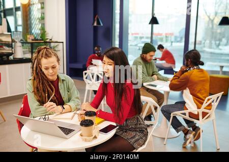 Two young contemporary intercultural businesswomen in casualwear working over presentation together while sitting by desk in office Stock Photo