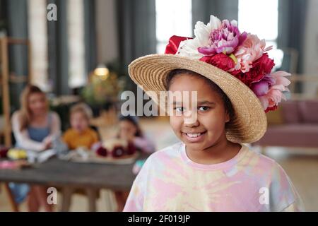 Pretty little mixed-race girl in hat decorated with large handmade flowers of white, pink and crimson colors standing against her friends by table Stock Photo