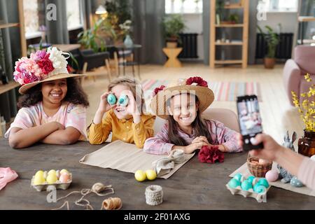 Three cheerful little children in casualwear looking at smartphone camera with toothy smiles while young female taking their photo by table Stock Photo