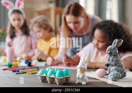 Grey and white toy rabbits and group of painted Easter eggs in egg-box standing on wooden table with kids drawing pictures on background Stock Photo