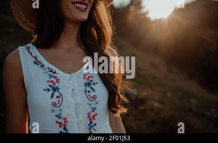 The midsection of a young smiling woman in light summer clothes wearing a hat letting her long hair shine under the sunset rays Stock Photo