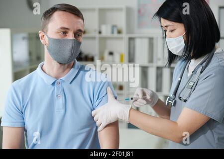 Female clinician or nurse of Asian ethnicity in uniform and protective mask vaccinating young man sitting in medical office of modern clinics Stock Photo