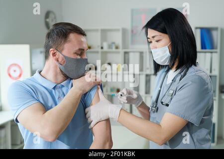 Female clinician or nurse of Asian ethnicity in uniform and protective mask making injection to young man sitting in front of her in medical office Stock Photo