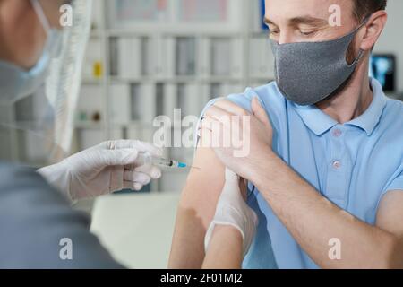 Young man holding sleeve of his shirt while nurse in protective workwear making him injection during vaccination against covid in clinics Stock Photo