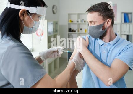 Asian nurse in uniform and protective workwear making injection to young man during vaccination against covid in contemporary clinics Stock Photo